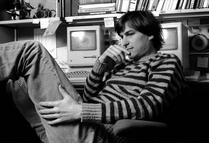 Steve Jobs in his office, leaning back in a chair beside an early Macintosh